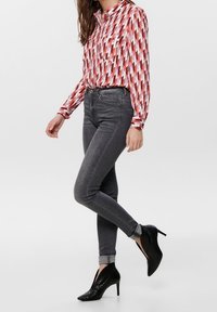 Blusa lunga a maniche con motivo rosso e rosa, abbinata a jeans skinny grigio scuro e stivaletti neri con tacco alto.