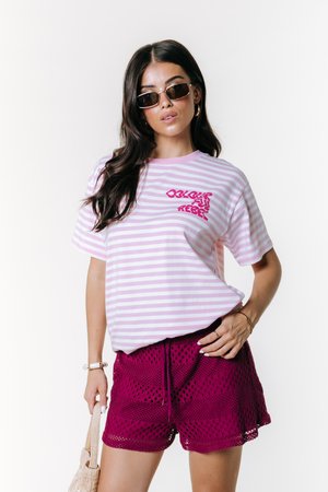 Maglietta a righe rosa e bianche con grafica "Colourful Rebel"; abbinata a pantaloncini viola all'uncinetto e occhiali da sole, mostrando un look estivo casual.