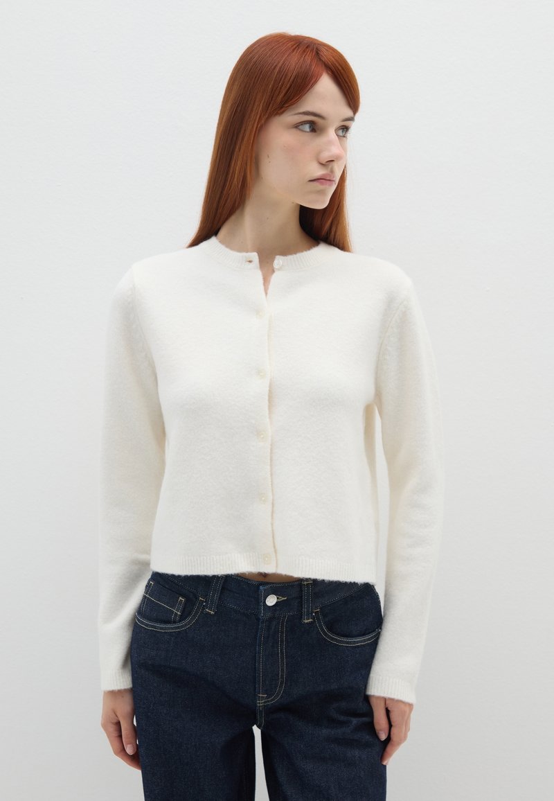 Cardigan bianco a maniche lunghe, con scollatura rotonda e bottoni frontali, realizzato in materiale morbido, abbinato a jeans di denim scuro.