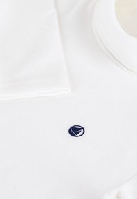 Hvit bomulls T-skjorte med rund hals, med en liten marineblå logo på forsiden og en jevn tekstur. Ermene er pent brettet.