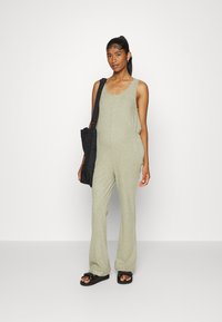 Macacão verde-oliva canelado com pernas largas, decote arredondado e bolsos laterais. Combinado com uma bolsa preta de ombro e sandálias rasas.