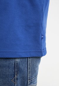 Blaues Baumwoll-T-Shirt mit geradem Saum, kurzen Ärmeln und einem kleinen Etikett an der Seite, kombiniert mit Denim-Jeans mit sichtbaren Nähten.
