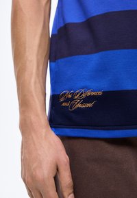 Άνδρας χέρι και χέρι δίπλα σε μπλε και μαύρη ριγέ μπλούζα με χρυσή κεντημένη φράση «Nos Différences nous Unissent» κοντά στο τελείωμα.