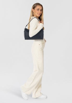 Bolso de mano de cuero azul marino oscuro con una textura suave, forma curvas y una sola asa. El modelo lleva una blusa de manga larga color crema y pantalones de pierna ancha.