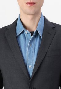 Dunkelgrauer Blazer mit glatter Textur, über einem hellblauen Denimhemd mit Knopfkraagen und sichtbaren Nähdetails geschichtet.