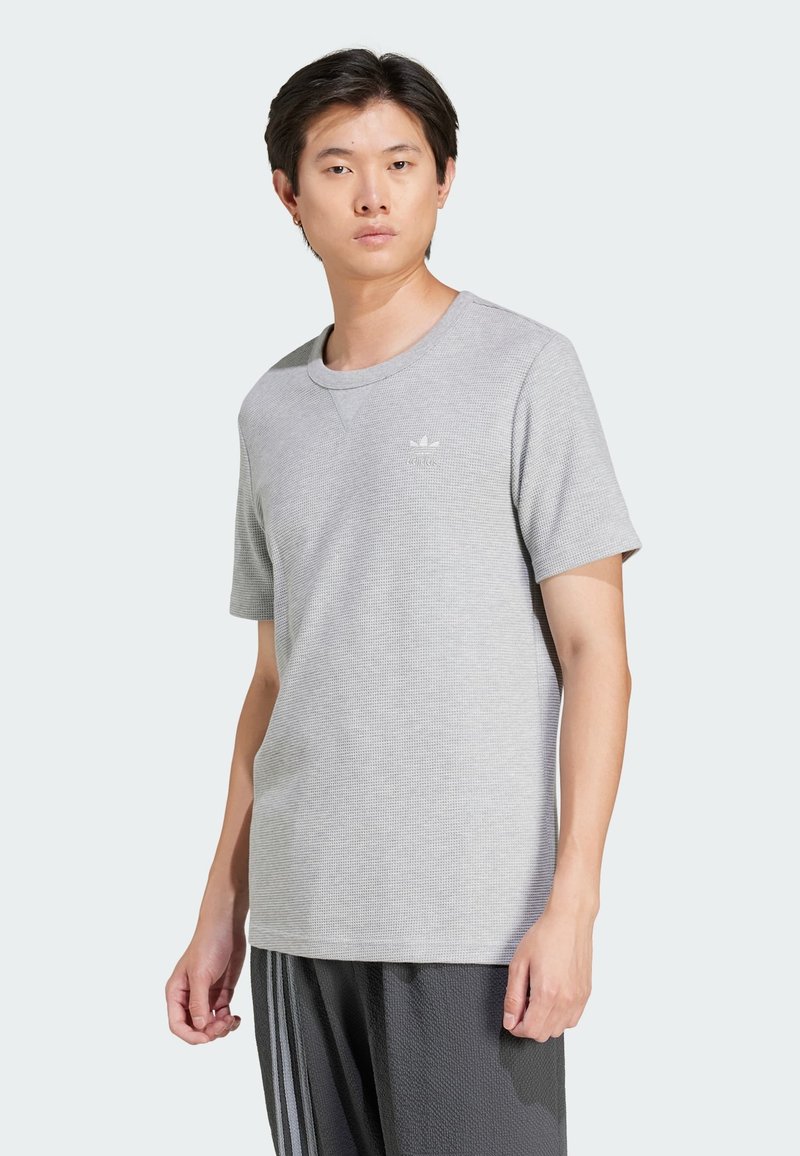 Šedé tričko s krátkym rukávom s textúrovanou tkaninou, okrúhlym výstrihom a malým bielym logom na ľavej hrudi. Skombinované s tmavými vzorovanými nohavicami.