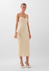 Beige midi-mekko, jossa on sydänleikkaus, ohuet hihnat, sileä pinta ja vartalonmyötäinen siluetti, yhdistettynä clutch-laukkuun ja korkokenkiin.