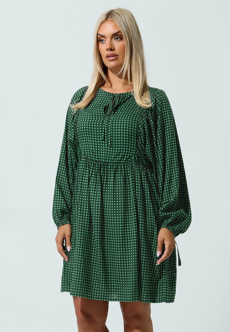 Nainen, jolla on pitkät vaaleat hiukset, pukeutuneena vihreään ruudulliseen pitkähihaiseen mekkoon, jossa on kootut vyötärö ja solmittava kaula-aukko, seisoo yksinkertaista taustaa vasten.