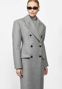 Szary dwurzędowy płaszcz wełniany z dużymi klapami, czarnymi guzikami i bocznymi kieszeniami. Model nosi szary golf i eleganckie ciemne okulary przeciwsłoneczne.