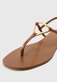 Sandalo thong marrone con strap sottili, dotato di un accento triangolare dorato. Suola piatta con una texture liscia e dettagli di cucitura minimi.