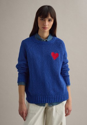 Donna che indossa un maglione blu lavorato a maglia con un cuore rosso sul petto sopra una camicia di jeans e pantaloni chiari, su uno sfondo semplice.