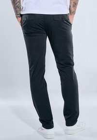 Black pants with a slim fit, featuring two back pockets with button closures, soft texture, and paired with white sneakers.