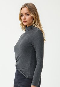 Blonde vrouw met blauwe ogen, gekleed in een donkergrijze lange mouwen top en zwarte broek, staande zijwaarts tegen een eenvoudige lichte achtergrond.