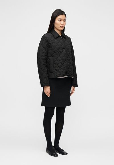 Μαύρο καπιτονέ μπουφάν με κουμπιά μπροστά, γιακά και πλαϊνές τσέπες, σε συνδυασμό με μαύρη φούστα και καλσόν, φορεμένο με μαύρα παπούτσια.