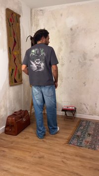 Άνδρας με ντραντλοκς σε φαρδιά τζιν και μπλούζα με γραφικό σχέδιο, στέκεται σε δωμάτιο με φθαρμένους λευκούς τοίχους, ένα χαλί και μια δερμάτινη τσάντα στο πάτωμα.