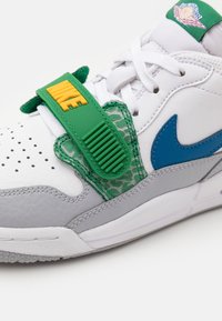 Bílé kožené tenisky s šedými akcenty, modrým swooshem, zeleným popruhem se žlutým textem "NIKE" a strukturovaným zeleným vzorem na popruhu.