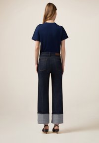 Tricou scurt cu mânecă, de culoare bleumarin, asortat cu blugi evazați din denim închis la culoare, cu talie înaltă, având manșete gri și o etichetă din piele maro pe spate.