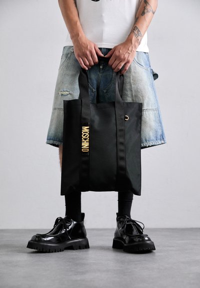Geantă neagră tip tote cu mânere duale, logo auriu "MOSCHINO", textură netedă și design minimal. Purttată de o persoană îmbrăcată în şorturi din denim și pantofi negri.