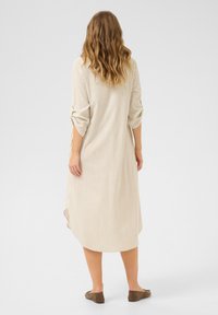 Beige pruhované šaty s dlouhými rukávy, knoflíkovými detaily na manžetách a asymetrickým lemem. Materiál vypadá lehce a měkce. Zobrazeny ploché boty.