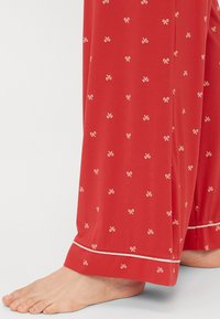 Pantalons de pyjama rouges avec de petits motifs de nœuds blancs, présentant une texture lisse et un ourlet blanc contrastant aux poignets.