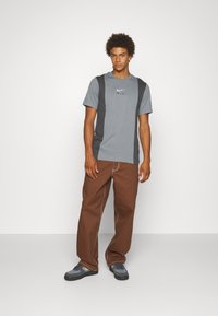 Grå t-shirt med svarta sidopaneler och en subtil Nike-logga, ihop med bruna lösa byxor och grå sneakers på en enfärgad bakgrund.