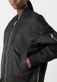 Schwarze Bomberjacke aus glattem Nylon mit einem Reißverschluss vorne, gerippten Bündchen, seitlichen Taschen und einer kleinen Tasche am Ärmel.