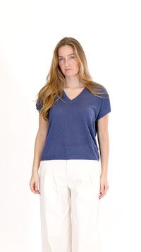 Žena se světlými dlouhými vlasy, oblečená v tmavě modrém tričku s krátkými rukávy a výstřihem do V a bílých širokých kalhotách s vysokým pasem, stojící na bílém pozadí.