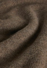 Detailný záber na mäkkú, hnedú pletenú tkaninu s viditeľnou textúrou stehov a jemnými záhybmi vytvárajúcimi nenápadné tiene.