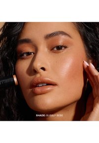 Nära bild av ett ansikte med en varmtonad, glansig hy som har highlighter. Nyans: Bubbly Bebe; naturlig läppfärg och mjuk textur.