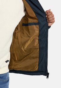 Tengerészkék dzseki belső része sima aranyszínű béléssel, oldalzsebbel és cipzáras záródással, kiemelve az ellentétes anyagokat és színeket.