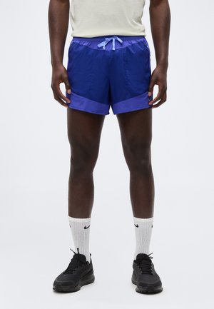 Atlet iført marineblå træningsshorts med lyseblå snor, hvide crew-strømper med sorte logoer og sorte løbesko, stående mod en hvid baggrund.