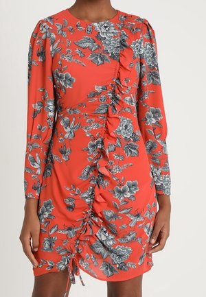 Rode jurk met lange mouwen, grijs bloemmotief en centrale geplooide ruches, gedragen door een staand persoon tegen een effen achtergrond.