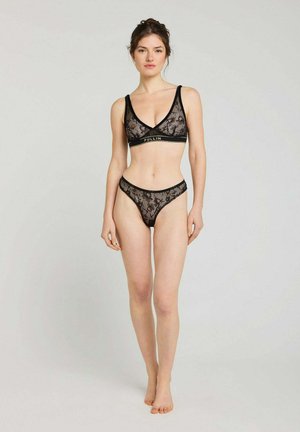 Femme debout portant un ensemble de lingerie en dentelle noire avec motif floral et bandes élastiques de marque sur fond clair uni.