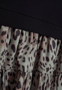 Schwarzes Oberteil mit geripptem Ausschnitt über einem plissierten Rock aus grünem Stoff mit braunem Leopardenmuster, das einen mehrschichtigen Look schafft.