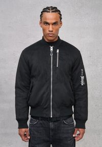 Giacca bomber nera con una texture liscia, chiusura frontale con zip, polsini a coste e dettaglio ricamato sulla manica. Indossata sopra una maglietta nera.