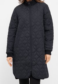Prošívaná černá bunda s vysokým límcem, předním zipem a bočními kapsami. Obsahuje diamantový vzor a žebrované manžety pro zvýšení textury.