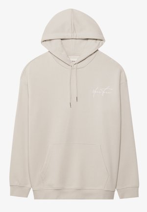 Beige huppari pehmeästä kankaasta, jossa on etupuolella kengurulaukku, kiristysnauha hupussa ja valkoinen kursivoitu logo vasemmassa rinnassa.