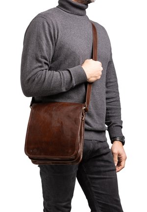 Uomo che indossa un dolcevita grigio scuro e pantaloni neri tiene una borsa a tracolla in pelle marrone incrociata sul petto.