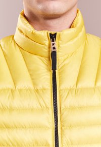 Gilet jaune matelassé avec col montant, fermeture éclair sur le devant et un petit logo "B" sur la tirette de la fermeture. Texture douce et lisse sans motifs supplémentaires.