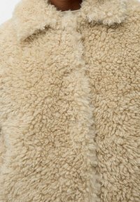 Beige falsk pelsjakke med en blød, struktureret overflade, der har en klassisk krave og knaplukning. Behageligt og varmt design til koldere vejr.