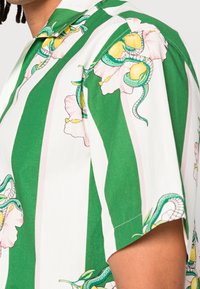 Košile s krátkými rukávy s proužkovaným vzorem v zelené a bílé barvě s motivy květin a hadů. Vyrobená z hladké látky s volným střihem.