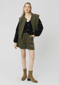 Gilet texturé vert sur blouse noire à manches longues à fleurs, associé à un short olive et des bottines beiges à talon carré.