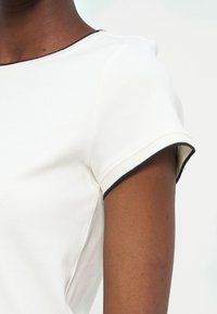 Κοντινό πλάνο ατόμου με σκούρο δέρμα που φοράει λευκό μπλουζάκι με κοντά μανίκια και μαύρο περίγραμμα στο λαιμό και στις άκρες των μανικιών.