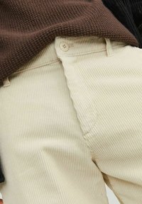 Pantaloni in corduroy crema con texture a coste prominenti, dotati di chiusura con bottoni e tasche laterali, abbinati a un maglione in maglia marrone.