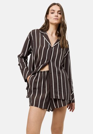 Vrouw draagt een zwart, lang mouwig, met knopen sluitend hemd en bijpassende korte broek met witte verticale strepen, staand met één hand in de zak.