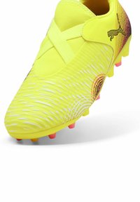Scarpa da calcio gialla brillante con upper testurizzato, cinturino incrociato e tacchetti colorati in rosa e arancione. Presenta un dettaglio con logo stilizzato.