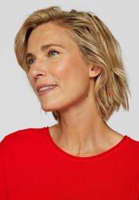 Rotes, geripptes Oberteil mit rundem Ausschnitt und kurzen Ärmeln. Das Modell hat kurzes, welliges blondes Haar und einen natürlichen Teint.