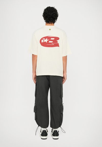 Maglietta oversize beige con un logo rosso sulla schiena, abbinata a pantaloni cargo larghi scuri e sneakers bianche e nere.