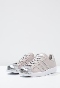 Sneaker in suede grigio chiaro con punta in metallo argento, suola in gomma bianca e strisce grigie ai lati. Design con lacci e collare imbottito.