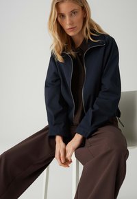 Femme blonde aux cheveux lâchés portant une veste noire à fermeture éclair et un pantalon large marron, assise sur un tabouret blanc devant un fond uni.